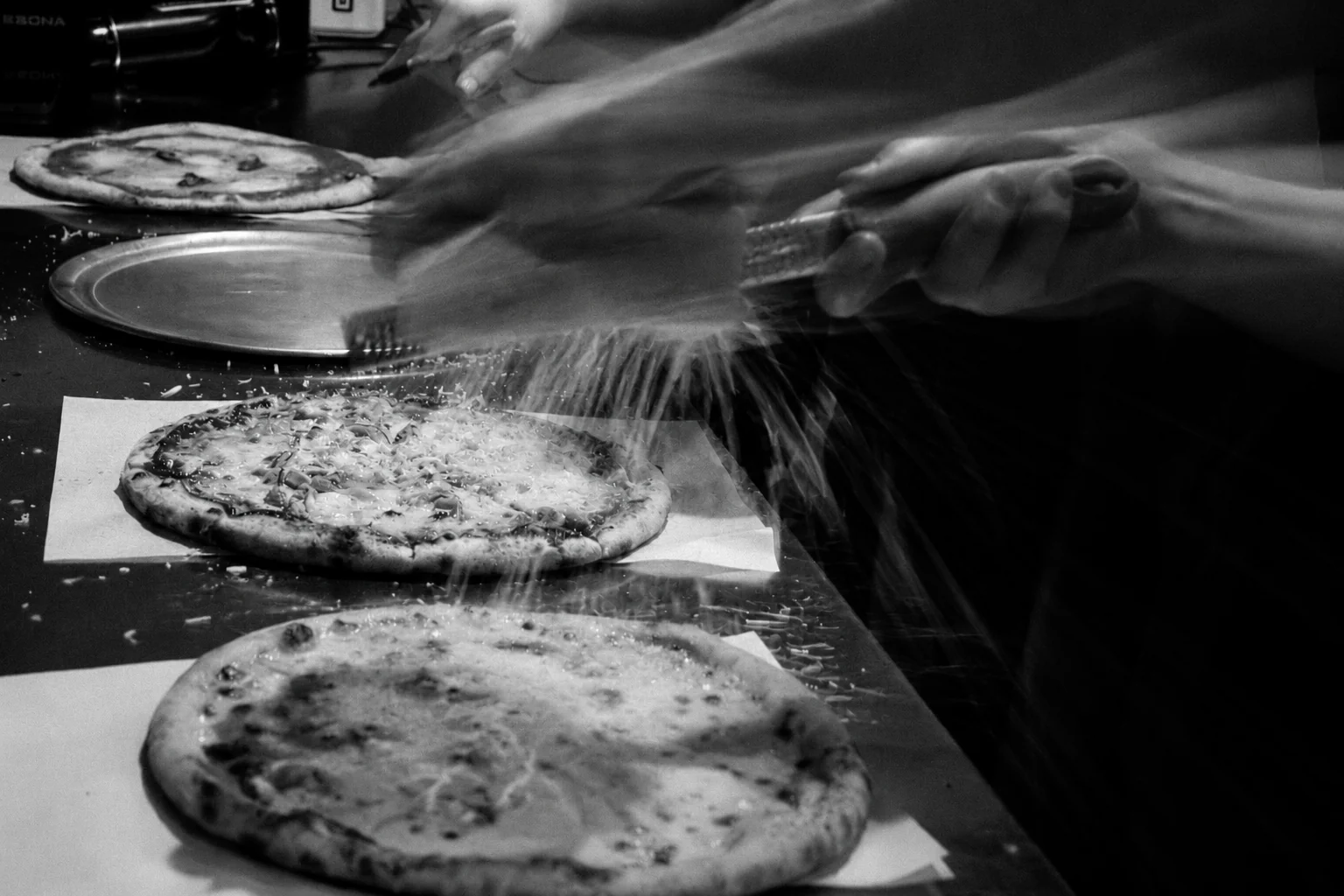 Cheese being grated over sourdough pizzas in black and white at Billy’s Pizza