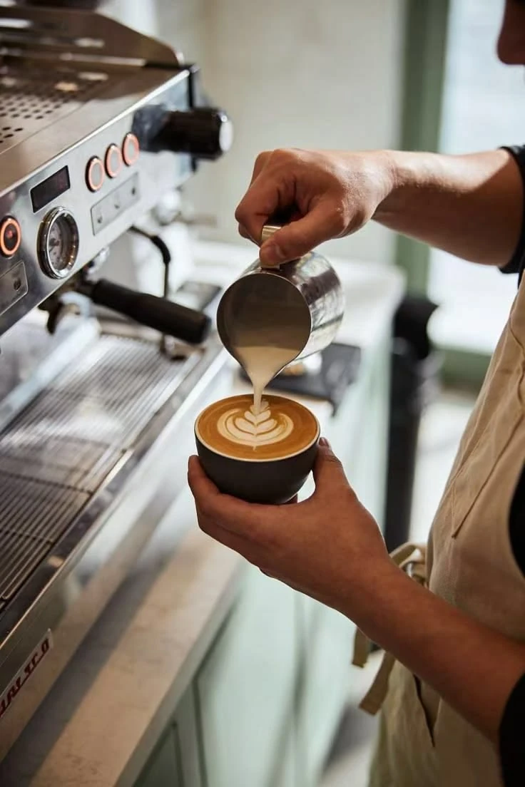 Barista pouring milk to create latte art beside an espresso machine